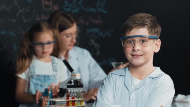 Smart boy crossing arms while teacher and friend doing experiment at STEM science class. Student mixing colored solution while standing at board written chemistry theory at laboratory. Erudition. - Footage, Video