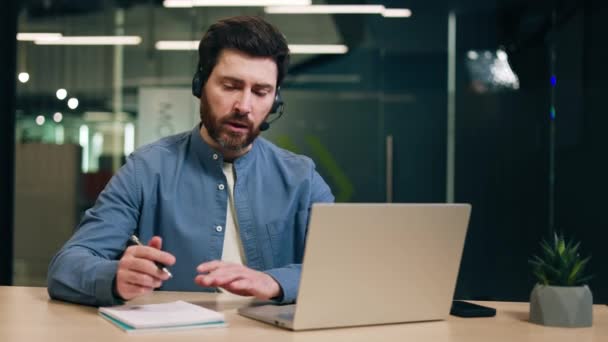 Hombre ocupado con auriculares inalámbricos y gestos con las manos mientras habla y mira la pantalla del ordenador portátil. Freelancer en camisa casual discutiendo sobre el trabajo durante una videollamada en línea en la computadora. - Imágenes, Vídeo
