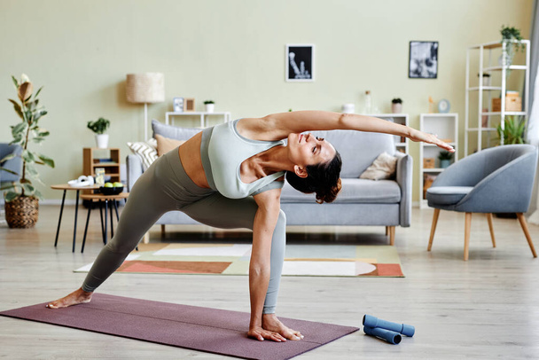 Porträt einer jungen Frau, die sich zu Hause dehnt, während sie mit geschlossenen Augen Yoga-Workout genießt - Foto, Bild