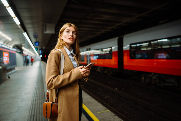 Hermosa mujer joven atractiva de pie en la plataforma de la estación en el tiempo de la noche, utilizando el teléfono inteligente, navegar por Internet o desplazarse a través de noticias  - Foto, imagen