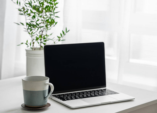 Schreibtisch mit Laptop, Kaffeetasse und Vase vor einem Fenster mit Morgenlicht. Gemütliches Bürokonzept im skandinavischen Stil. - Foto, Bild