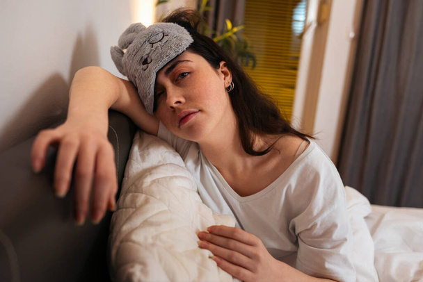 Κουρασμένη λευκή γυναίκα που φοράει μάσκα ύπνου και κάθεται στο κρεβάτι με μαξιλάρι. Έννοια της αϋπνίας και αϋπνίας. - Φωτογραφία, εικόνα