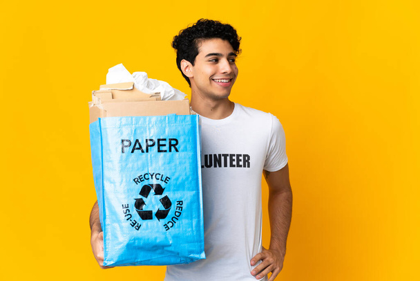 Νεαρός Βενεζουέλας κρατώντας μια τσάντα ανακύκλωσης γεμάτη χαρτί για ανακύκλωση ποζάροντας με τα χέρια στο γοφό και χαμογελώντας - Φωτογραφία, εικόνα