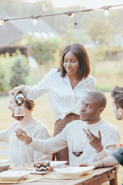 На фотографії зображена чорношкіра афроамериканка, яка уважно виливає вино в келих гостей під час багаторазової расової вечері. Сцена відбувається в теплій, сяючій атмосфері, як - Фото, зображення