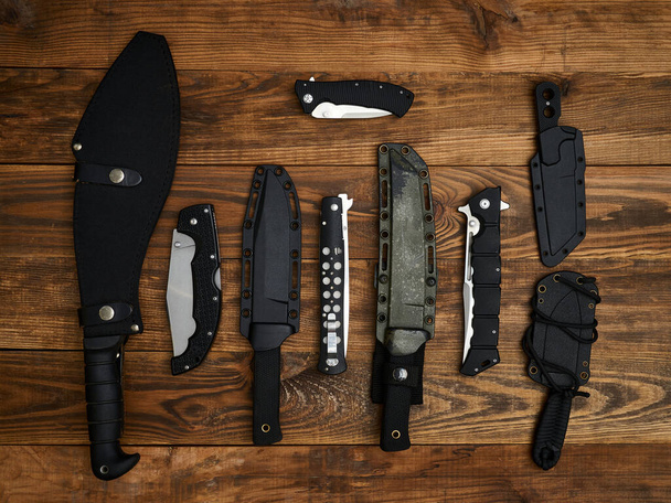 Nahaufnahme von neun verschiedenen Messern mit geschlossenen Klingen oder in Scheiden. Alles ausgebreitet auf dem braunen Holzgrund. Festklinge, Taschenmesser und taktische Messer. Schwarze Griffe. - Foto, Bild