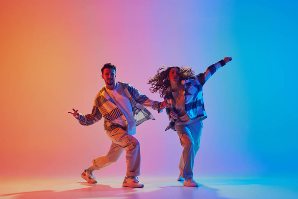 Casal dançante se move em sincronia, homem e mulher, dançando em estúdio iluminado por néon contra fundo gradiente. Expressão energética. Estilo urbano. Conceito de movimento, energia, batalhas de dança. Retrato de gel dinâmico - Foto, Imagem