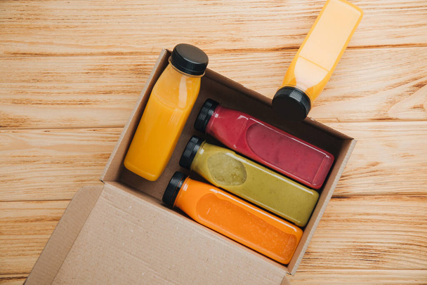 Hedelmät ja vihannekset smoothiet läpinäkyvissä muovipulloissa. Punainen, vihreä, oranssi smoothies pahvilaatikossa puupöydällä. Terveellisen ruokavalion käsite. Yläkuva - Valokuva, kuva