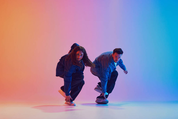 Dva tanečníci provádějící synchronní hip-hop pohyby oblečeni v džínových šatech na pozadí gradientu v neonovém světle. Koncept kultury mládeže, hudby, životního stylu, stylu a módy, akce. Gelový portrét. - Fotografie, Obrázek
