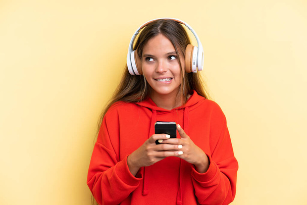 Junge kaukasische Frau isoliert auf gelbem Hintergrund Musik hören mit einem Handy und Denken - Foto, Bild