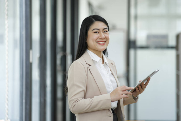 Профессиональная и уверенная в себе деловая женщина, улыбающаяся в камеру, стоящая у своего стола с ноутбуком в современном офисе. - Фото, изображение