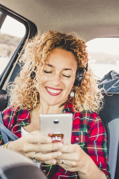 Alegre mujer feliz sentarse en los asientos traseros dentro de un coche moderno utilizando un teléfono celular para escuchar música con auriculares - personas que disfrutan de viajar con el concepto de tecnología - Foto, Imagen