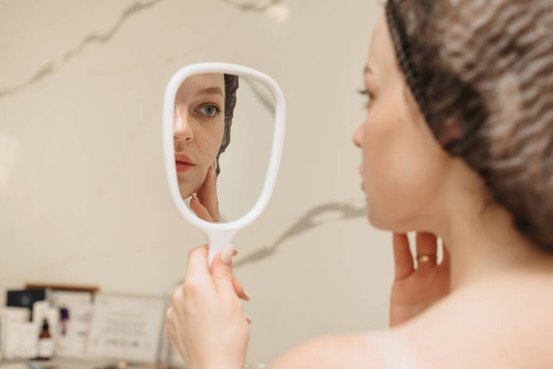 Una mujer sonriente se mira en el espejo en la oficina del cosmetólogo - Foto, Imagen