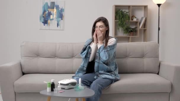 Młoda kobieta w ubraniu casual, przy użyciu aerozolu do nosa w celu ułatwienia oddychania, siedzi na kanapie w domu - Materiał filmowy, wideo