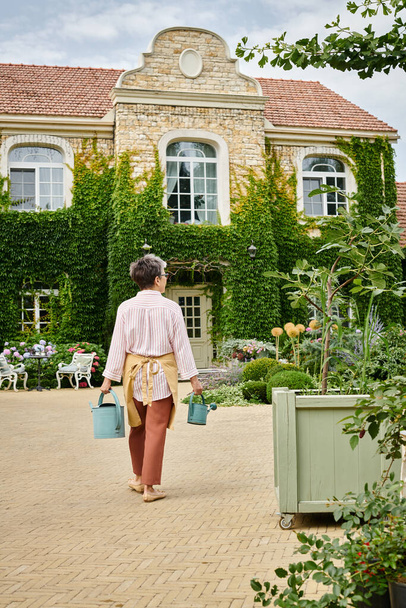 イギリスの彼女の家の前に2つの水やり缶を保持する居心地の良い服装の成熟した女性のバックビュー - 写真・画像