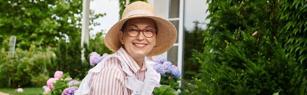 atractiva mujer madura alegre con sombrero cuidando de su hortensia y sonriendo a la cámara, pancarta - Foto, Imagen