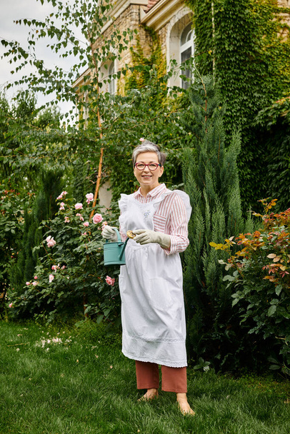 attractive mature jolly woman with glasses holding watering can and smiling happily at camera - Foto, Imagem