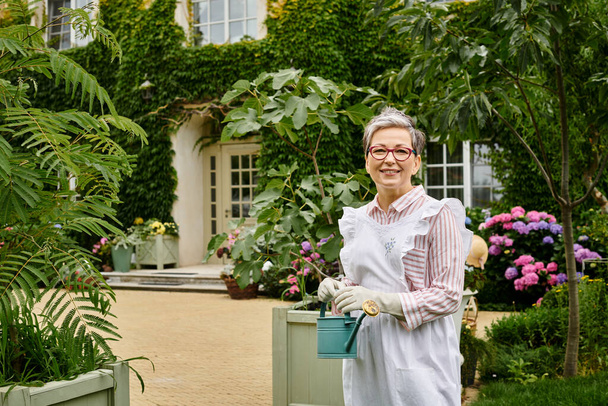 attractive mature jolly woman with glasses holding watering can and smiling happily at camera - Photo, Image