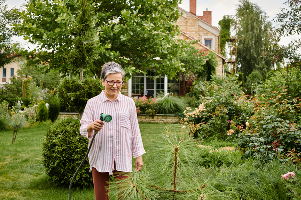 όμορφη χαρούμενη ώριμη γυναίκα με γυαλιά χρησιμοποιώντας μάνικα για να ποτίσει τα ζωντανά φυτά της στον κήπο της - Φωτογραφία, εικόνα