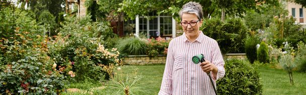 beautiful jolly mature woman with glasses using hose to water lively plants in her garden, banner - Foto, imagen