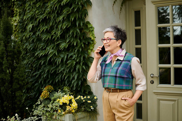 mature cheerful elegant woman with glasses and short hair talking by her phone near house in England - Фото, изображение