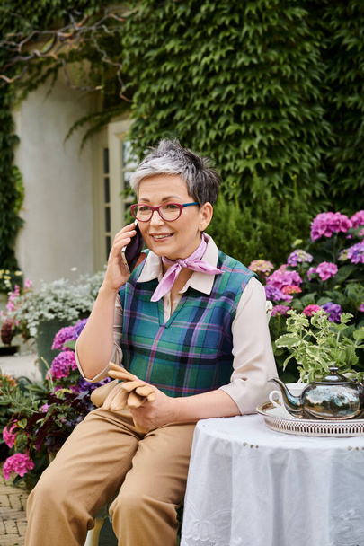 dojrzała pogodna kobieta siedzi w ogrodzie w czasie herbaty i rozmawia przez telefon w pobliżu domu w Anglii - Zdjęcie, obraz