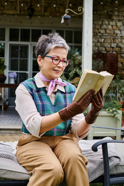 sophisticated mature joyous woman with glasses reading book near her house in rural England - Photo, Image