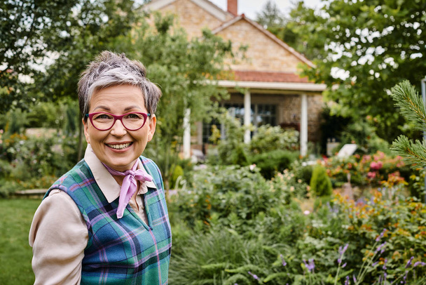 kultivierte reife fröhliche Frau mit kurzen Haaren, die in der Nähe ihres Hauses in England in die Kamera lächelt - Foto, Bild