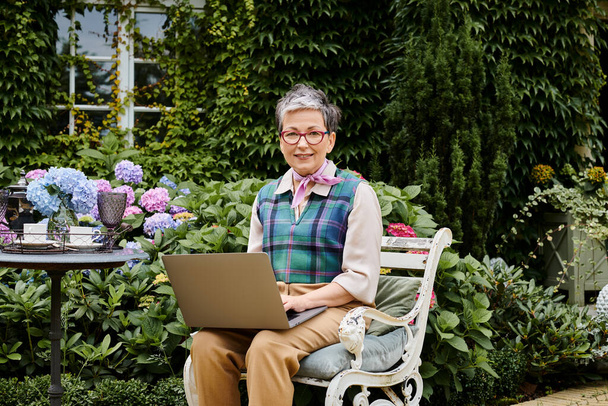 élégante femme mature gaie avec les cheveux courts assis avec ordinateur portable à l'heure du thé et souriant à la caméra - Photo, image