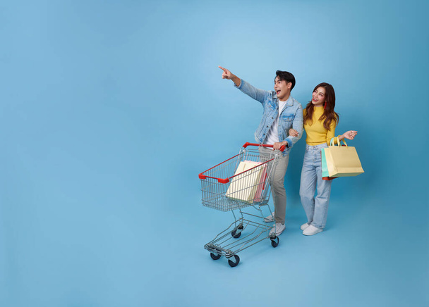Full size jovem alegre casal asiático se divertir carro carro de carrinho de compras e segurando compras saco de compras supermercado no shopping isolado no fundo azul - Foto, Imagem