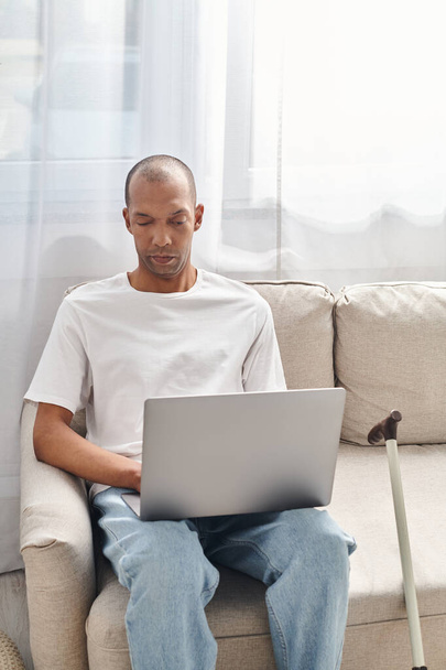 Un uomo con miastenia grave usa un computer portatile su un divano, vicino al bastone da passeggio - Foto, immagini