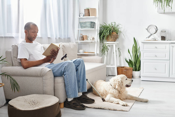 Een Afro-Amerikaanse man met myasthenia gravis zit op een bank, een boek te lezen, vergezeld van zijn trouwe Labrador hond. - Foto, afbeelding