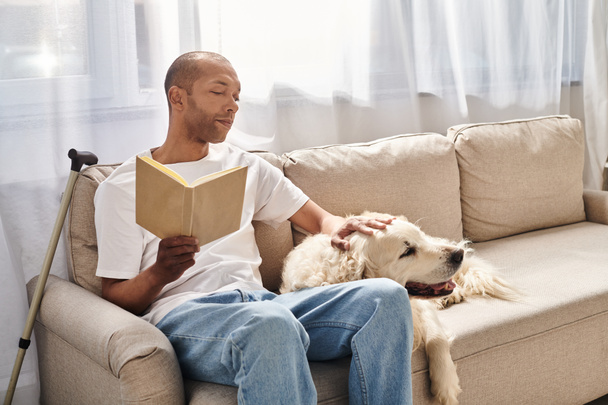 Niepełnosprawny Afroamerykanin z miastenią Gravis czytający książkę na kanapie obok swojego lojalnego psa Labradora. - Zdjęcie, obraz