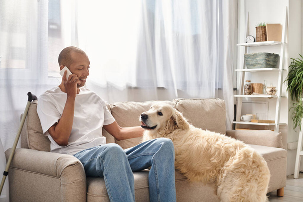 Een Afro-Amerikaanse man, gehandicapt met myasthenia gravis, zit op een bank te praten op een mobiele telefoon naast zijn trouwe Labrador hond. - Foto, afbeelding