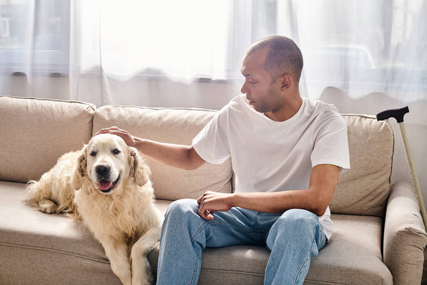 A man, disabled with myasthenia gravis, sits on a sofa petting a Labrador dog, showcasing diversity and inclusion. - Фото, изображение