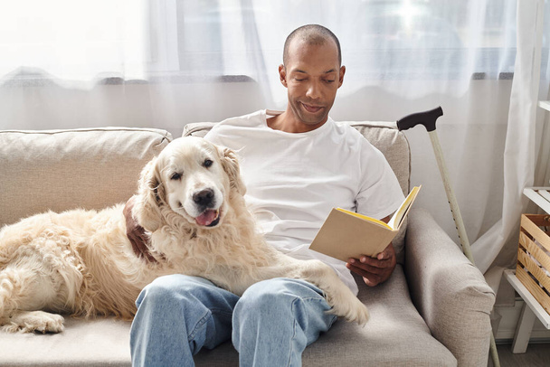 Ένας άνδρας με μυασθένεια Gravis χαλαρώνει στο σπίτι σε έναν καναπέ με πιστό σκυλί Λαμπραντόρ του, απορροφημένος σε ένα καλό βιβλίο. - Φωτογραφία, εικόνα