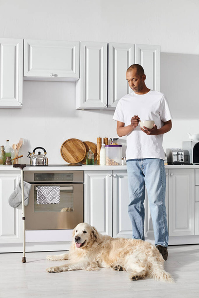Инвалид афроамериканец с миастенией и грависом стоит на кухне рядом со своей верной собакой-лабрадором. - Фото, изображение