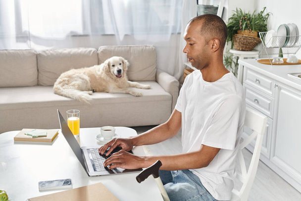 障害のあるアフリカ系アメリカ人男性がラブラドール犬を伴うラップトップコンピュータを使用してテーブルに座ります.. - 写真・画像
