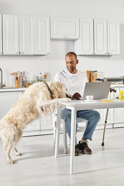 Een gehandicapte Afro-Amerikaanse man zit aan een tafel met een laptop voor zich open, begeleid door zijn trouwe Labrador hond. - Foto, afbeelding