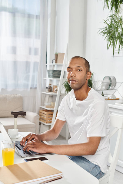 Ein Mann afroamerikanischer Abstammung, der mit Myasthenia gravis lebt, benutzt an einem Tisch einen Laptop. - Foto, Bild