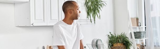 Gehandicapte Afro-Amerikaanse man met myasthenia gravis syndroom staat in de keuken naast een potplant. - Foto, afbeelding
