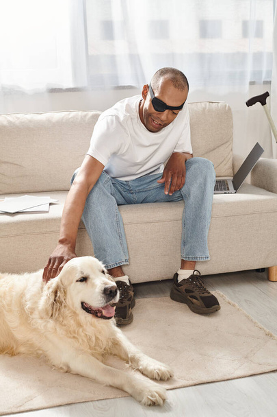 Egy mozgássérült afro-amerikai férfi myasthenia gravis szindrómában ül egy hűséges labrador kutya mellett a kanapén.. - Fotó, kép
