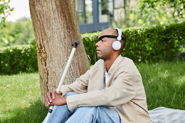 Um homem com fones de ouvido está sentado em um cobertor ao lado de uma árvore, desfrutando de música e do ambiente tranquilo - Foto, Imagem