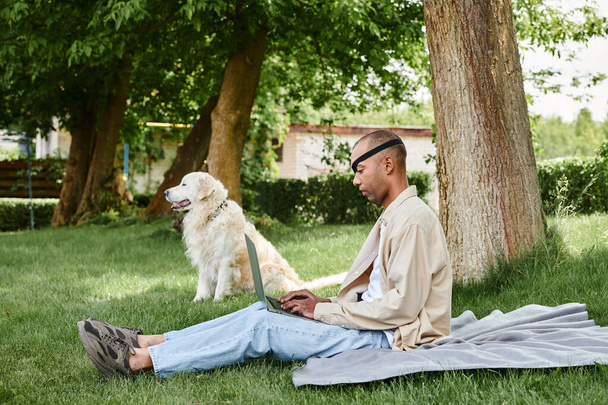 Разнообразный человек с Миастенией Гравис сидит на траве, используя ноутбук в сопровождении своей верной собаки-лабрадора. - Фото, изображение