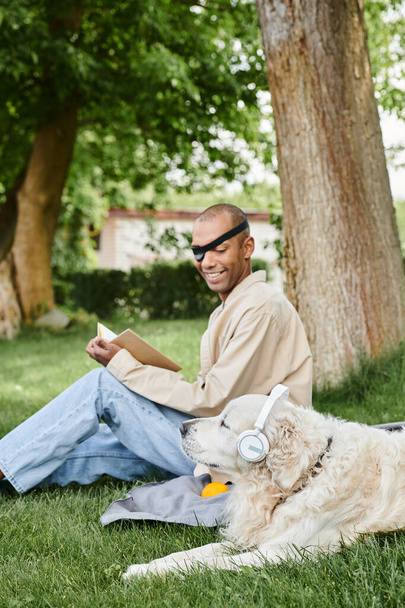 Miyastenia gravis sendromu olan bir adam labrador köpeğiyle çimenlerde oturuyor, ikisi de kulaklık takıyor.. - Fotoğraf, Görsel