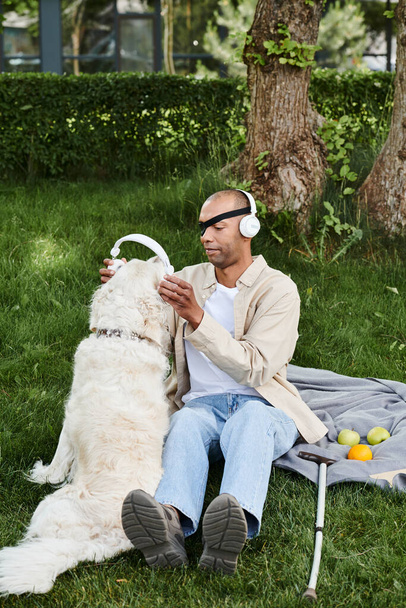 Un hombre afroamericano con miastenia gravis se sienta con su perro labrador en la hierba, ambos disfrutando de la música a través de auriculares. - Foto, Imagen