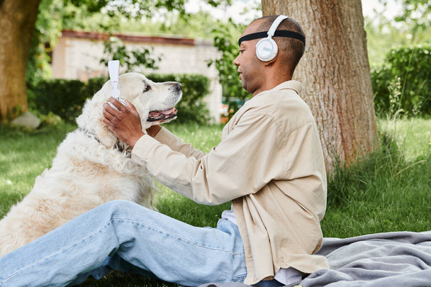 Un uomo afroamericano disabile con la sindrome della miastenia grave siede nell'erba con un cane Labrador che indossa le cuffie, godendo di un momento di pace insieme. - Foto, immagini