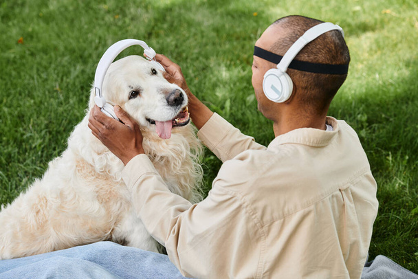Un uomo diverso con la sindrome della miastenia grave siede sull'erba, accarezzando gioiosamente il suo fedele cane Labrador mentre entrambi indossano le cuffie. - Foto, immagini