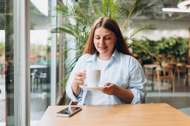 Mujer feliz usar camisa azul sostener taza beber café sentarse solo en la mesa en la cafetería, cafetería o restaurante en el interior. Concepto de desayuno fácil - Foto, Imagen
