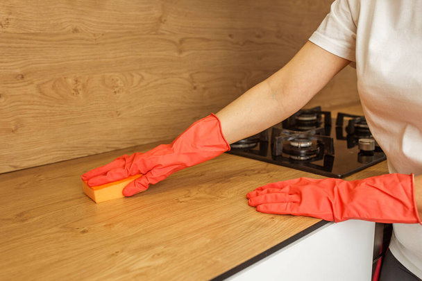 Κοντινό πλάνο των χεριών του ατόμου σε κόκκινα γάντια καθαρισμού μια ξύλινη επιφάνεια κουζίνας με σφουγγάρι και πορτοκαλί σπρέι καθαρισμού. - Φωτογραφία, εικόνα