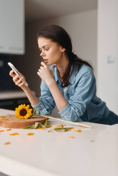 Mladá běloška sedí doma, drží smartphone a čte si textovou zprávu u stolu v kuchyni.. - Fotografie, Obrázek